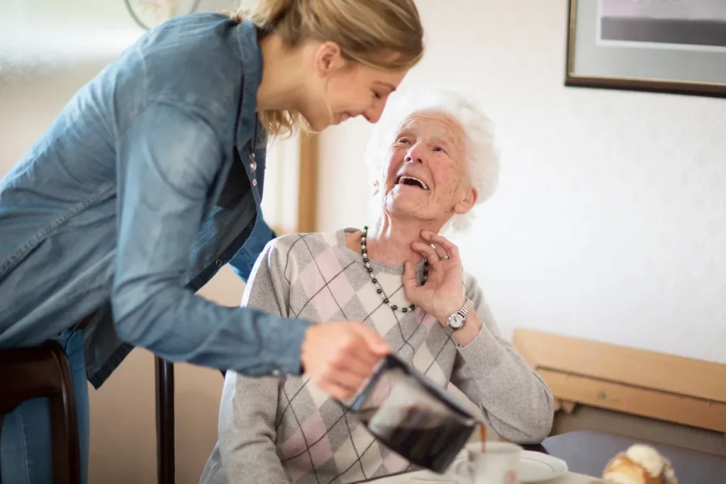 A smiling young woman pours coffee for an elderly woman at a table. The elderly woman, wearing a diamond-patterned sweater, looks up joyfully. Cozy atmosphere.