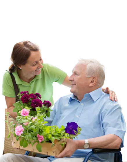 A woman in a green shirt smiles warmly at an elderly man in a wheelchair, holding a wooden box of vibrant flowers. The background is black, highlighting their joyful interaction.
